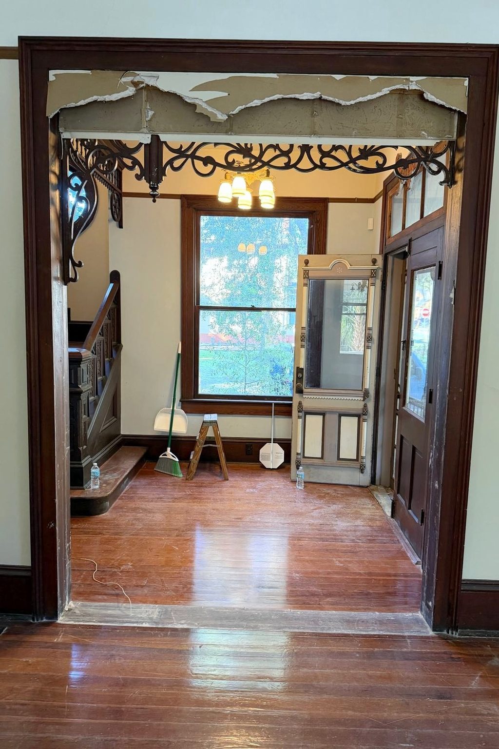 Today: the entry hall seen through a wide doorway, mid-work, with torn drywall above the fretwork band, a ladder, a broom and a loose screen door.