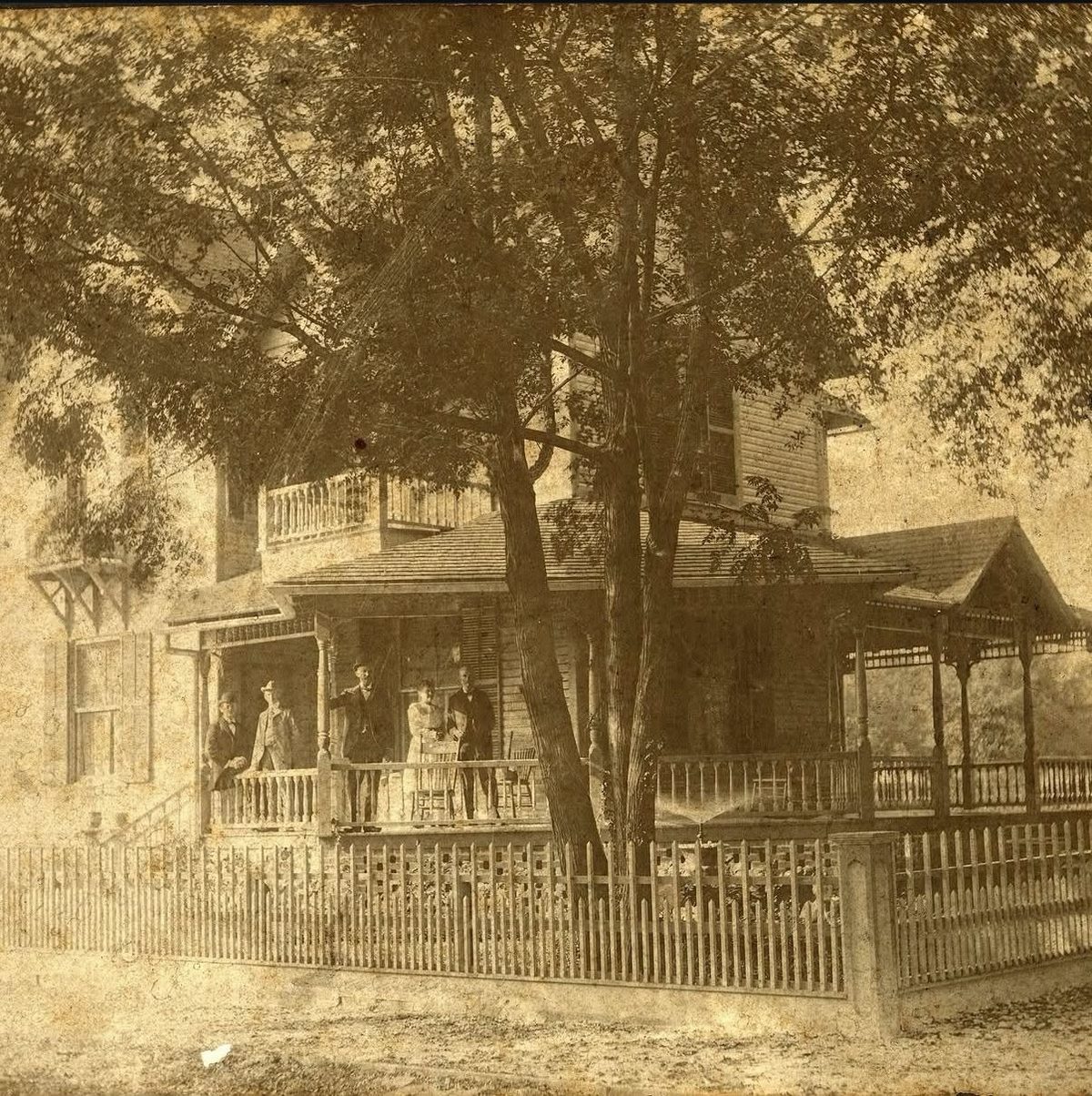 A sepia photograph of the house in its early years, with five people in period dress on the wraparound porch behind a picket fence.