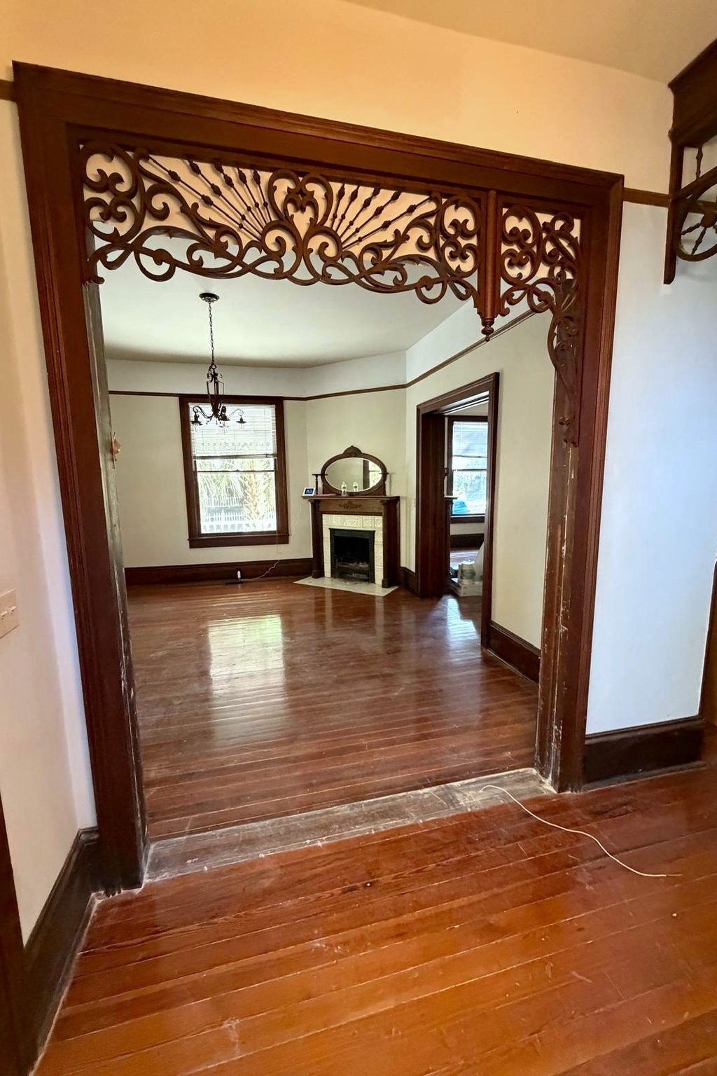 Today: the living room seen through the carved fretwork arch, empty, with wood floors, a mirrored mantel and a chandelier.