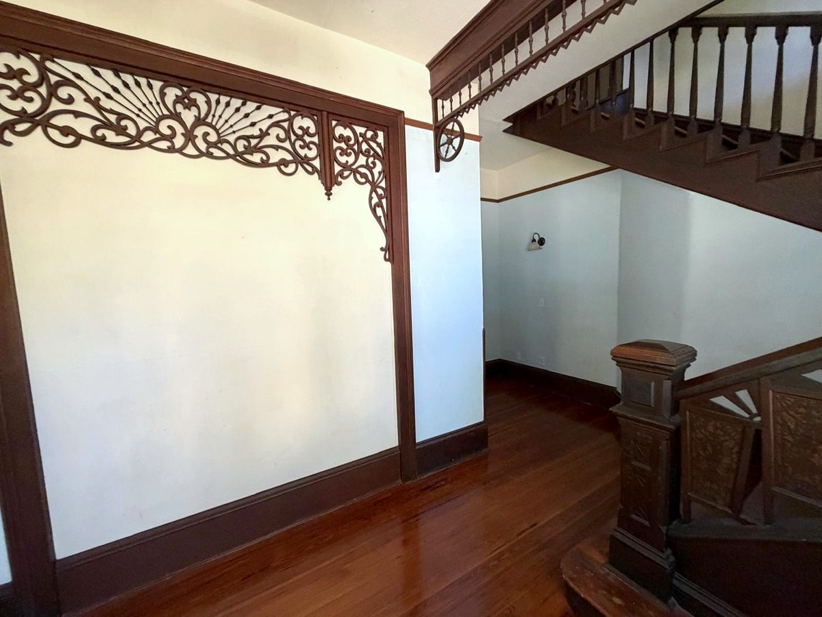 The stair hall: a fretwork arch on the left, a spindled frieze with wheel brackets overhead, and the carved newel post at the foot of the stairs.