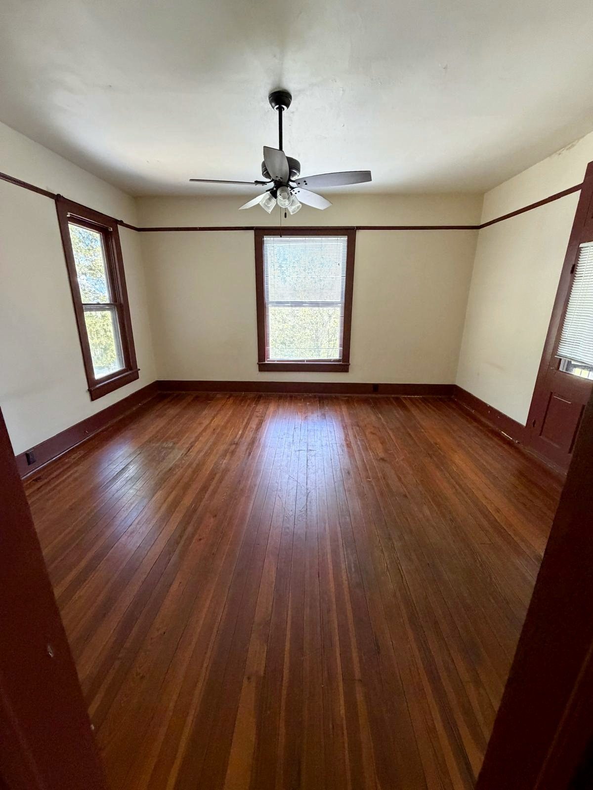 An empty upstairs room with three tall windows looking into the trees, a ceiling fan, dark-stained trim and wood floors.