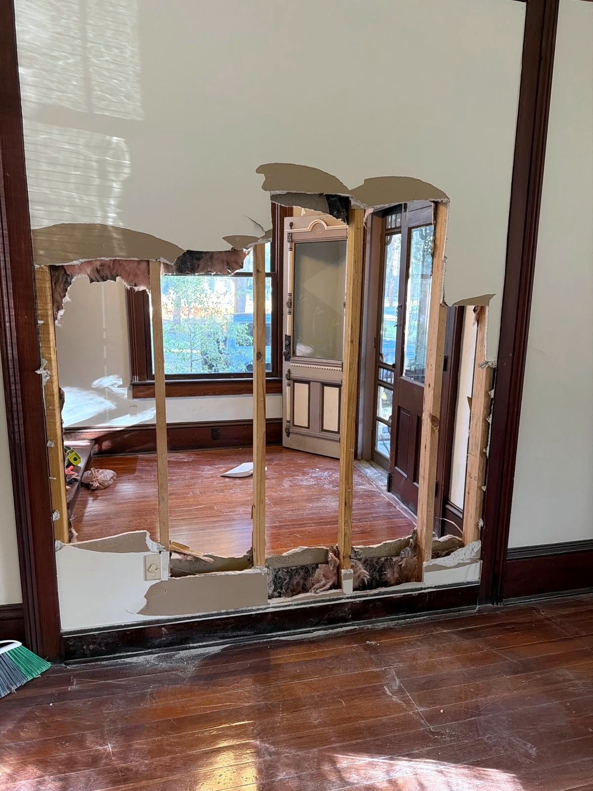 A wall opened down to the studs, looking through to the entry hall with its tall window and front doors.