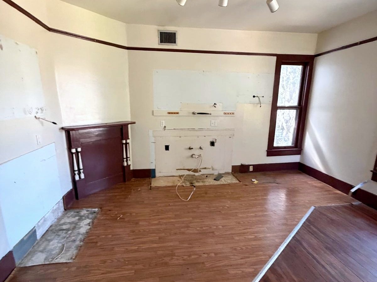 The same kitchen wall with the cabinets, counters and appliances removed, leaving plumbing stubs, patched plaster and the painted mantel.